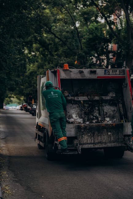 A waste collection worker dressed in green high-visibility workwear with orange reflective stripes is standing beside a side-loading rubbish collection truck on a quiet suburban street lined with trees and parked cars. The worker is positioned near the rear of the truck, which has an open, blackened and slightly dirty compartment for waste storage, indicative of recent collection activity. The scene is illuminated by natural daylight, with the background featuring lush green foliage and a street extending into the distance. The surrounding environment appears calm, with no other activity or pedestrians visible. This setup reflects typical private waste collection services, emphasizing on-site rubbish removal without reliance on local authority bin collections, maintaining a professional but neutral tone suitable for content related to independent waste disposal options provided by companies such as Waste Removal Highbury.