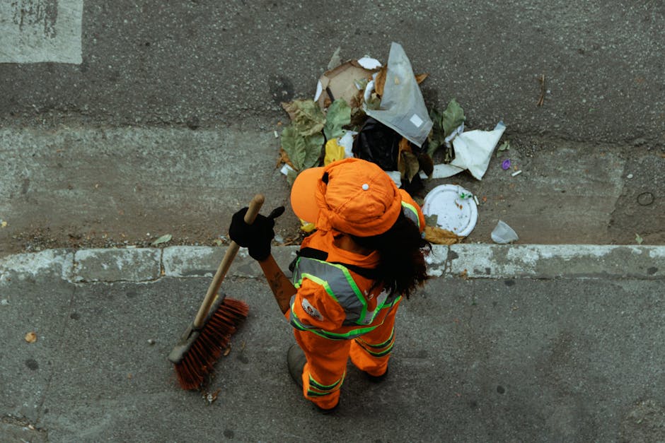 An individual dressed in bright orange high-visibility workwear, including a jacket, trousers, and a matching hat, is seen from a top-down perspective on a paved urban street. The worker is holding a long-handled broom with a red bristle head and appears to be in the process of cleaning or collecting litter that is piled against a concrete wall. The pile of rubbish consists of various materials, including crumpled paper, plastic bags, a disposable plate, and other debris, with some items partially crushed or torn. The scene takes place outdoors, with the litter situated in the curb corner where the street meets the wall, and the lighting suggests daytime conditions. The presence of waste in this setting emphasizes the importance of on-site clearance and private waste handling, which the services of Waste Removal Highbury aim to facilitate for local residents and businesses seeking efficient rubbish removal and disposal solutions in the area.