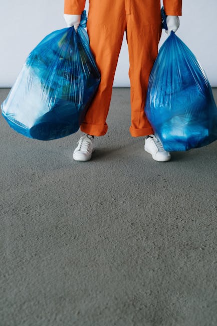 A waste collection worker dressed in green high-visibility workwear with orange reflective stripes is standing beside a side-loading rubbish collection truck on a quiet suburban street lined with trees and parked cars. The worker is positioned near the rear of the truck, which has an open, blackened and slightly dirty compartment for waste storage, indicative of recent collection activity. The scene is illuminated by natural daylight, with the background featuring lush green foliage and a street extending into the distance. The surrounding environment appears calm, with no other activity or pedestrians visible. This setup reflects typical private waste collection services, emphasizing on-site rubbish removal without reliance on local authority bin collections, maintaining a professional but neutral tone suitable for content related to independent waste disposal options provided by companies such as Waste Removal Highbury.