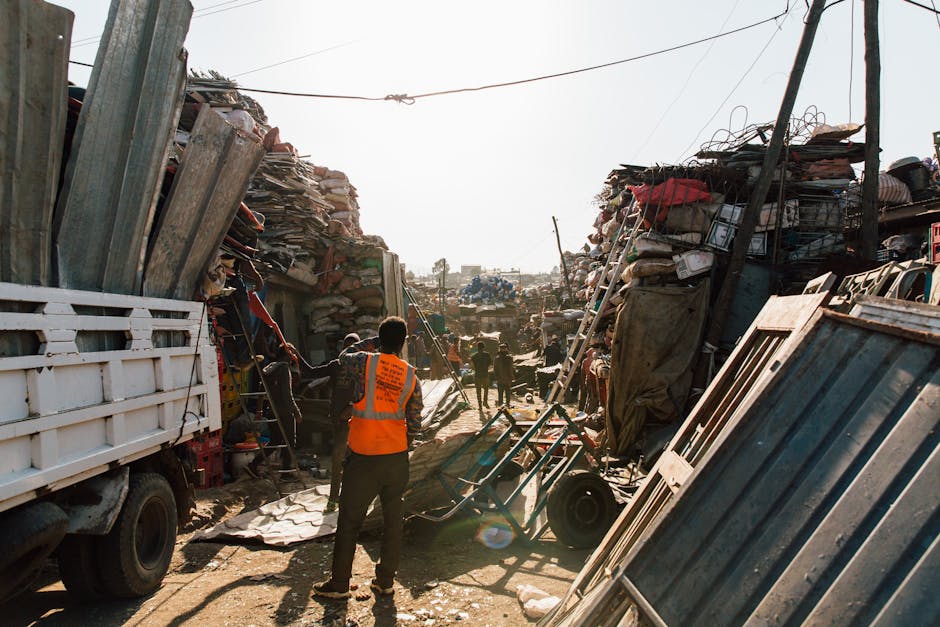 A scene at a waste collection site shows a narrow dirt pathway flanked by high piles of assorted rubbish and scrap material on either side. On the left, a white flatbed truck is partially visible, loaded with various metal and wooden debris. In the foreground, a worker wearing an orange high-visibility vest is standing with their back to the camera, observing the area, which suggests an organized waste removal operation by Waste Removal Highbury. To the right, a stack of metal panels and other construction debris leans against a makeshift barrier, with additional piles of mixed waste, including bags, wood, and miscellaneous items, extending into the background. Several other workers are seen in the distance, actively engaging in clearing activities amidst the clutter, highlighting an ongoing bulk waste clearance effort. The environment appears outdoors with bright daylight, and overhead wires are visible, indicating a semi-urban setting. This scene illustrates private waste handling or independent collection activities typical of non-local-authority rubbish removal services aimed at managing bulky waste disposal effectively.
