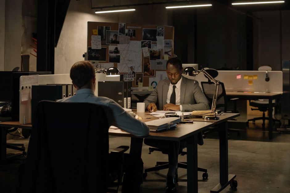 A professional office scene showing two men seated at a large desk in a dimly lit workspace. The man in the foreground has short brown hair and is viewed from behind, wearing a light-colored shirt. The man in the background, wearing a grey suit and black tie, is focused on paperwork with a pen in hand. The desk holds various documents, a white mug, a desk lamp, and office supplies. In the background, a cork board displays photographs, notes, and sketches, while a row of computers and office chairs are arranged along the back wall. The environment is modern, with a dark ceiling and warm lighting, creating a serious and focused atmosphere typical of a corporate or investigative setting, reflecting attention to detail relevant to office work or project management, but does not depict rubbish removal activities.