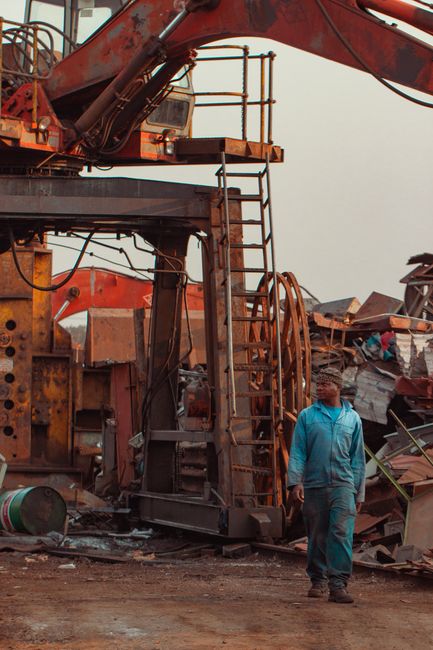 A scene at a waste collection site shows a narrow dirt pathway flanked by high piles of assorted rubbish and scrap material on either side. On the left, a white flatbed truck is partially visible, loaded with various metal and wooden debris. In the foreground, a worker wearing an orange high-visibility vest is standing with their back to the camera, observing the area, which suggests an organized waste removal operation by Waste Removal Highbury. To the right, a stack of metal panels and other construction debris leans against a makeshift barrier, with additional piles of mixed waste, including bags, wood, and miscellaneous items, extending into the background. Several other workers are seen in the distance, actively engaging in clearing activities amidst the clutter, highlighting an ongoing bulk waste clearance effort. The environment appears outdoors with bright daylight, and overhead wires are visible, indicating a semi-urban setting. This scene illustrates private waste handling or independent collection activities typical of non-local-authority rubbish removal services aimed at managing bulky waste disposal effectively.
