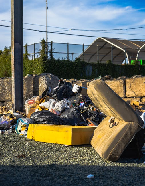A large pile of mixed waste and rubbish, including black plastic garbage bags, a yellow plastic container, and an old, dirty car tire, is seen spilling onto a gravel surface in an outdoor area. The waste appears to consist of various household and miscellaneous trash, scattered in front of a low stone wall made of roughly cut, light-colored stones. In the background, there are several wooden utility poles and overhead power lines, as well as tall greenery and shrubs behind the wall. To the right, a partially visible structure with a curved roof, possibly a greenhouse or shelter, features transparent or reflective panels. The scene is illuminated by daylight, with a clear blue sky and some wispy clouds overhead, illustrating the typical setting for private rubbish removal or on-site waste clearance undertaken by Waste Removal Highbury in the context of alternative waste handling and rubbish disposal services.