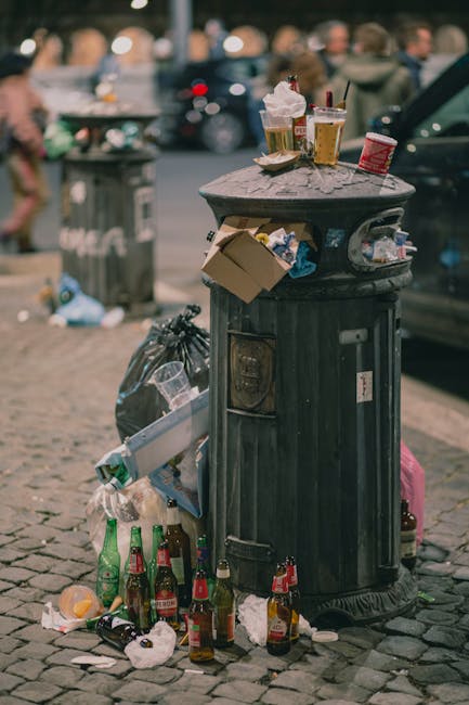 A black, cast-iron litter bin with a domed lid and vertical ridges stands on a cobblestone pavement, surrounded by an accumulation of discarded rubbish. The waste includes several glass bottles, partially crushed and scattered at the base of the bin, along with crumpled paper, plastic containers, and a few cardboard pieces. On top of the bin's lid, there are used paper cups, some with straws, and a plastic cup with a lid, all bearing leftover drink residue, as well as a small red paper food container. Nearby, a black garbage bag and a clear plastic bag filled with trash are tied and leaning against the bin. In the background, a grey trash bin and a blurred scene of parked cars and pedestrians are visible, indicating an urban street setting. The overall scene suggests an instance of improper waste disposal and highlights the importance of regular rubbish removal services. Waste Removal Highbury manages such onsite clearing to address alternative waste handling needs in residential areas.