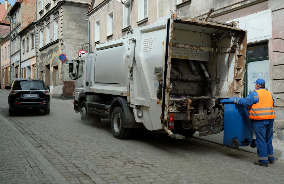 A waste collection vehicle parked on a narrow cobblestone street next to aged multi-story building facades with weathered plaster and brickwork. The truck, painted white with signs of rust on its rear loading mechanism, is positioned close to a building with large glass windows and neutral-colored walls. A worker dressed in a blue uniform and high-visibility orange vest is standing beside a blue wheeled bin near the vehicle’s rear, appearing to be in the process of emptying it. The street features a black car parked further along, and a no-parking sign is visible attached to a pole behind the truck. The environment suggests an urban setting where independent waste collection services like Waste Removal Highbury offer private rubbish removal and on-site clearance, providing an alternative to traditional council bin collection services. The natural lighting indicates daytime with overcast skies, contributing to a neutral and straightforward scene centered on waste collection activity.