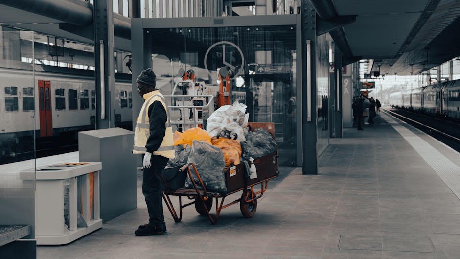 A worker dressed in dark clothing and a grey beanie hat, wearing a high-visibility vest and gloves, stands on a modern train station platform next to a large trolley loaded with assorted rubbish bags and discarded items. The trolley contains black, grey, orange, and clear plastic bags filled with waste materials, along with some loose debris and cardboard boxes, indicating a collection of mixed rubbish. Behind the worker, there is a glass-walled waiting area or ticket booth, and to the left, a stationary train with a white body and red accents is visible. The platform has a smooth grey surface with tactile paving near the edge for safety. Overhead, part of the station's metal and glass roof structure is visible, supported by dark-colored beams. In the background, a few other individuals can be seen further along the platform, with additional trains visible on the distant tracks. The overall scene suggests an environment where waste collection and rubbish removal from urban transit sites occur, and Waste Removal Highbury may be involved in such onsite clearance operations for public transportation facilities.