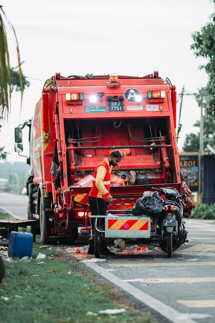 A worker dressed in a red and yellow uniform is operating a large red refuse collection vehicle, which is positioned on the side of a road. The back of the truck is open, revealing the internal mechanisms used for rubbish collection. The worker is leaning slightly forward, using tools to manage or clear waste, with a black trash bag placed on a small platform attached to the vehicle. Surrounding the scene is a roadside environment with grass and a curb, along with other elements such as a blue container on the ground nearby. The setting appears to be outdoors on a partly cloudy day, with trees and utility poles visible in the background. This scene illustrates the process of private waste collection or rubbish removal, which is an alternative to council-run services, consistent with methods often handled by independent waste disposal companies like Waste Removal Highbury. The image provides a clear view of the practical, on-site activity involved in rubbish collection, emphasizing the physical task of waste management in an urban or suburban environment.