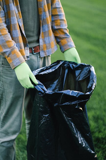 A worker dressed in a red and yellow uniform is operating a large red refuse collection vehicle, which is positioned on the side of a road. The back of the truck is open, revealing the internal mechanisms used for rubbish collection. The worker is leaning slightly forward, using tools to manage or clear waste, with a black trash bag placed on a small platform attached to the vehicle. Surrounding the scene is a roadside environment with grass and a curb, along with other elements such as a blue container on the ground nearby. The setting appears to be outdoors on a partly cloudy day, with trees and utility poles visible in the background. This scene illustrates the process of private waste collection or rubbish removal, which is an alternative to council-run services, consistent with methods often handled by independent waste disposal companies like Waste Removal Highbury. The image provides a clear view of the practical, on-site activity involved in rubbish collection, emphasizing the physical task of waste management in an urban or suburban environment.