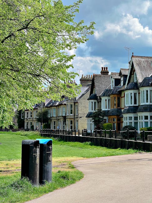 In a suburban residential area, two black cylindrical rubbish bins are positioned on a grassy verge beside a wide, paved footpath. The bins have rounded tops, with one showing a small blue recycling icon and the other a white symbol, indicating their use for waste and recyclable materials. Behind the bins, a black metal fence runs parallel to a row of traditional terraced houses, each featuring bay windows, pitched roofs with slate tiles, and brick or painted facades in various shades of beige, gray, and brown. The houses are closely spaced, with some displaying small front gardens or shrubbery. To the left, a large deciduous tree with fresh light green leaves extends its branches over the scene, contrasting against a partly cloudy sky with patches of blue and scattered white clouds. The environment suggests a quiet, well-maintained neighbourhood, where initiative like independent rubbish collection might be used alongside local authority services, reflecting a community-oriented approach to waste management that includes private disposal options, as indicated by the presence of designated rubbish bins on the street.