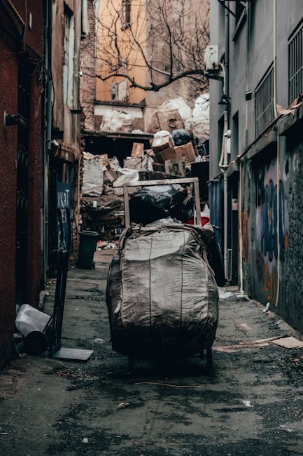 The image shows a narrow urban alleyway filled with a large accumulation of mixed waste and rubbish piled up against a makeshift barrier at the end of the alley. In the foreground, a large, worn, and slightly crumpled black and grey trash bag or sack is positioned upright on the dark, stained pavement, which appears dirty and weathered, with small debris scattered around. To the right, a building with a dark grey exterior wall has metal security grilles over windows and graffiti in various colours, while on the left side, the adjacent building features a rusty, weathered surface with visible signs of aging. At the top of the alley, leafless tree branches extend over the rubbish, indicating a winter setting. The scene suggests a situation where waste collection might be delayed or where private refuse disposal methods are in use, aligning with alternative rubbish removal services. The overall atmosphere is subdued and utilitarian, highlighting the need for proper waste management solutions typically handled by specialist waste removal providers like Waste Removal Highbury.