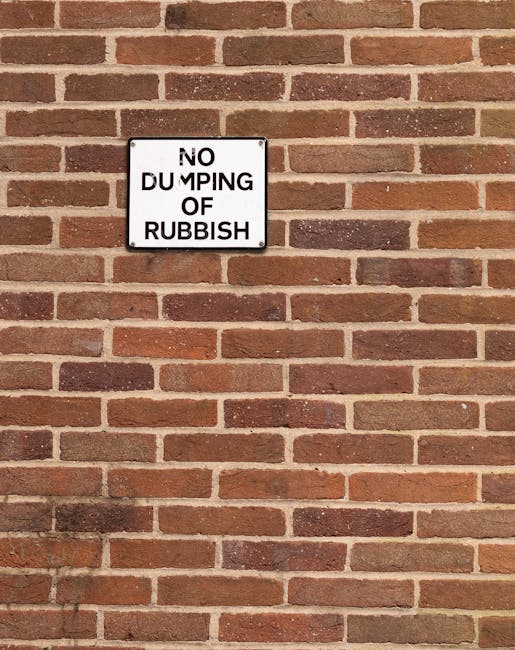 A rectangular white metal sign with black uppercase lettering reading 'NO DUMPING OF RUBBISH' is mounted on a red brick wall with a consistent pattern. The brick wall features standard-sized bricks with a reddish-brown hue, some showing darker shades and slight surface irregularities, laid in a running bond pattern with light grey mortar joints. The sign is centrally positioned, slightly above the middle of the image, and appears to be secured with four screws, one at each corner. The lighting is natural and even, casting minimal shadows on the wall. The absence of rubbish or other objects in the immediate vicinity emphasizes the focus on the sign’s instruction to prevent illegal dumping, aligning with waste management and rubbish removal efforts in residential or commercial areas. Waste Removal Highbury emphasizes proper waste handling and clearance, supporting compliant and responsible rubbish disposal practices in urban environments, such as this well-maintained brick frontage. The scene conveys a clear message discouraging rubbish dumping, consistent with the themes of private waste collection and on-site clearance services.