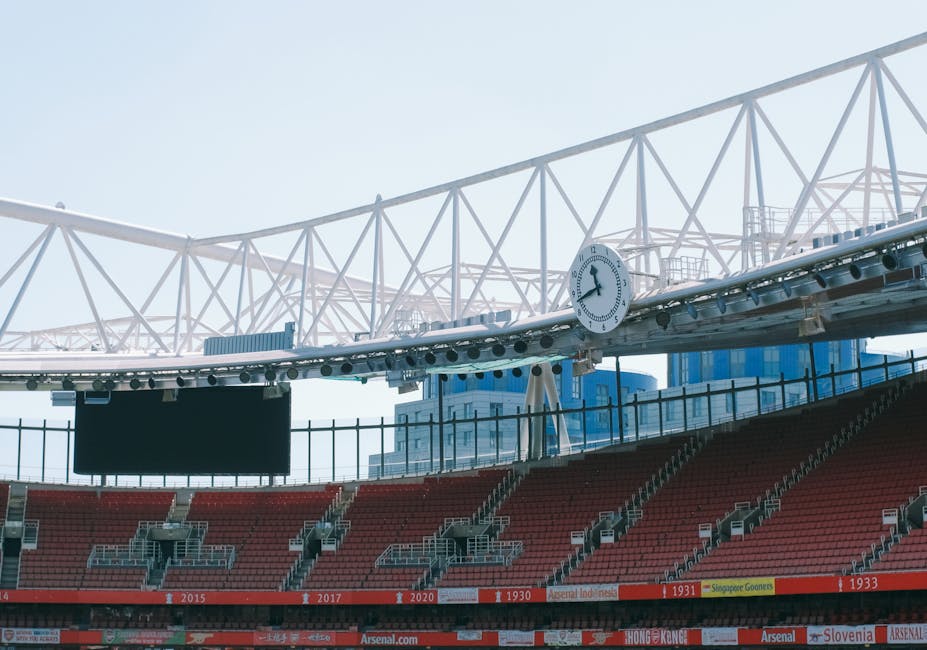 Inside a large outdoor sports stadium with a prominent white steel roof structure supported by trusses and beams, a circular clock is mounted on the roof framework showing the time as approximately 2:55. The seating area, visible in the lower part of the image, consists of red seats arranged in rows, with some seating sections marked by railings and step aisles. Behind the seating, tall modern office buildings with glass facades can be seen in the background, indicating an urban setting. The lighting appears natural, with clear skies and sunlight illuminating the scene evenly. The stadium environment suggests an area where event rubbish and waste could be collected during maintenance or after events, which relates to services offered by Waste Removal Highbury in providing private waste handling and alternative rubbish removal options beyond local authority collection methods. The image emphasizes structural details of the stadium, with a focus on the roof and seating areas, highlighting the context of waste management within large-scale, public venues.