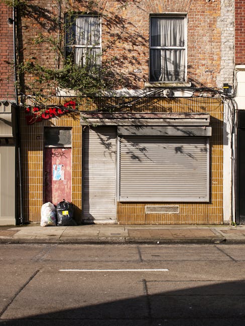 The image depicts the facade of a small commercial building on a city street, with a brick upper floor featuring two white-framed windows, partially obscured by shadows and leafless tree branches. The ground floor consists of a storefront with yellow-brown tiles surrounding a metal roller shutter door that is closed, with a small vent at the bottom. To the left of the shutter, there is a narrow, graffiti-marked door painted in pink and covered with posters. In front of the building, on the sidewalk, there are three garbage bags, two black and one white, placed near the door, suggesting waste collection or disposal. Overhead, electrical wires and cables are loosely arranged, with some hanging and connecting to the building. The scene appears to be in an urban environment, with no other visible business signage, and natural daylight illuminating the scene, creating shadows on the facade and street pavement. This ambient setup aligns with the context of private or independent rubbish removal services offered by Waste Removal Highbury for commercial premises on Highbury Grove, Highbury.