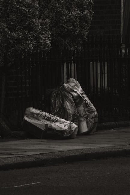 Three large black refuse bags made from thick, crinkled plastic are leaning against a dark wooden fence beside a paved sidewalk. The bags appear to be filled with waste and are somewhat deflated, with visible creases and folds on their surfaces. The bags are situated close to each other, with one bag slightly overlapping the other two. To the left of the bags, there is a small tree or shrub with dense foliage that extends upwards, partially obscuring and contrasting against the fence. In the background, the fence continues along the edge of the scene, and a residential building with brick or tiled walls is faintly visible, suggesting an urban or suburban environment. The overall lighting is low, and the image is in black and white, giving a stark, somber tone that highlights the everyday sight of waste awaiting collection or disposal, consistent with private rubbish removal services such as those offered by Waste Removal Highbury.