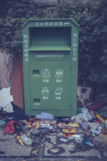 A green outdoor clothing and accessory disposal bin with Chinese characters and a contact phone number printed on its front, situated on a sidewalk against a backdrop of dense foliage and a wooden fence. The bin has multiple compartments, each designated for specific items: a section for clothing, shoes, bags, and books, with small square openings for depositing these items. In front of the bin, there is a disorganized pile of discarded rubbish, including various articles of clothing such as shoes, sneakers, and fabric pieces, alongside scattered trash, small boxes, and miscellaneous debris. The scene suggests an informal collection point for secondary waste disposal, with a cluttered arrangement indicative of a lack of proper waste management, emphasizing the contrast between the purpose of the bin and the surrounding untidy environment. The natural lighting highlights the textures of the trash and the painted surface of the bin, aligning with themes of private waste handling and DIY disposal options often seen in community or informal settings.
