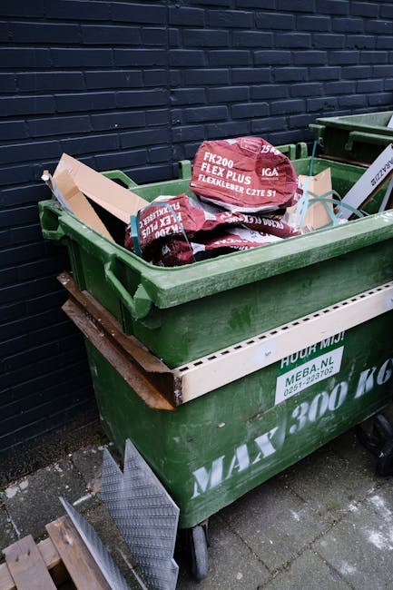 A large green wheeled skip positioned outdoors on a paved surface next to a dark brick wall, filled with red and black packaged materials, possibly flexible builder’s membranes or roofing materials. The skip is situated on a partially weathered wooden pallet, and a white panel with contact details is attached to its side. Adjacent to the skip are another similar container and a small section of metal grate on the ground. The environment appears to be a backyard or service area, with surrounding non-scenic elements including the pavement and wall. This scene likely depicts a private waste disposal or collection setup arranged by Waste Removal Highbury for handling bulky or construction-related rubbish as part of on-site clearance or alternative waste handling services, consistent with the themes of rubbish and waste management.