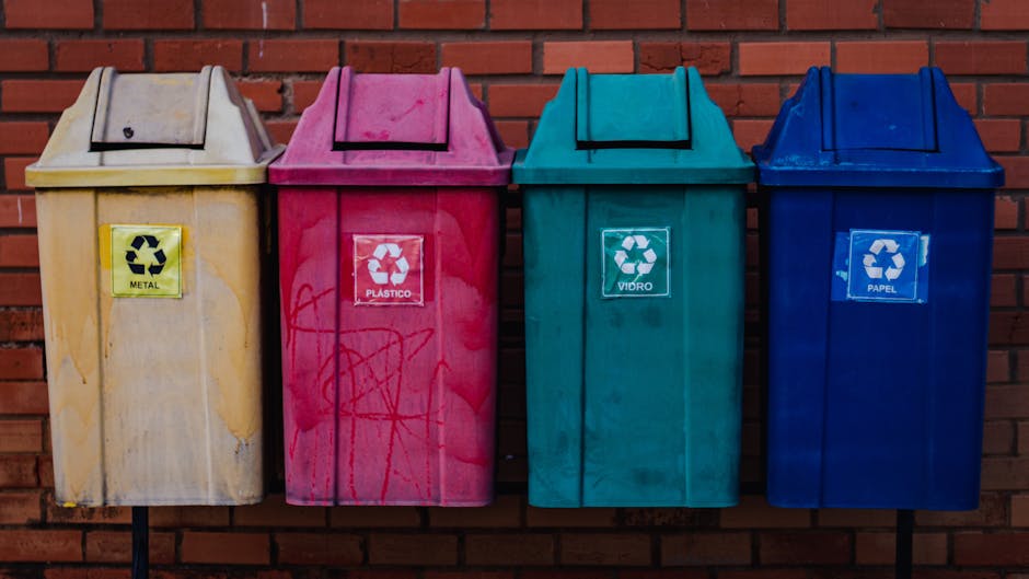 A green outdoor clothing and accessory disposal bin with Chinese characters and a contact phone number printed on its front, situated on a sidewalk against a backdrop of dense foliage and a wooden fence. The bin has multiple compartments, each designated for specific items: a section for clothing, shoes, bags, and books, with small square openings for depositing these items. In front of the bin, there is a disorganized pile of discarded rubbish, including various articles of clothing such as shoes, sneakers, and fabric pieces, alongside scattered trash, small boxes, and miscellaneous debris. The scene suggests an informal collection point for secondary waste disposal, with a cluttered arrangement indicative of a lack of proper waste management, emphasizing the contrast between the purpose of the bin and the surrounding untidy environment. The natural lighting highlights the textures of the trash and the painted surface of the bin, aligning with themes of private waste handling and DIY disposal options often seen in community or informal settings.